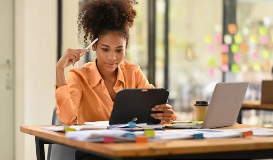 Businesswoman in an orange shirt working at a desk, holding a tablet and pen while reviewing documents with a laptop and office supplies nearby.