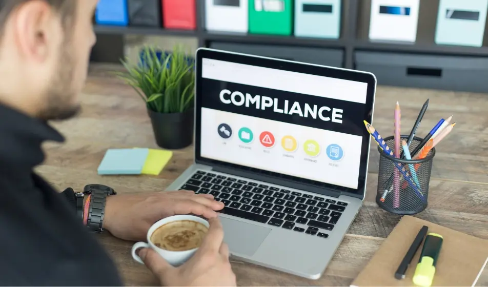 Man in the corner of the screen, back facing us, holding a coffee - his laptop is in front of him and reads 'compliance' on the screen in bold capital letters