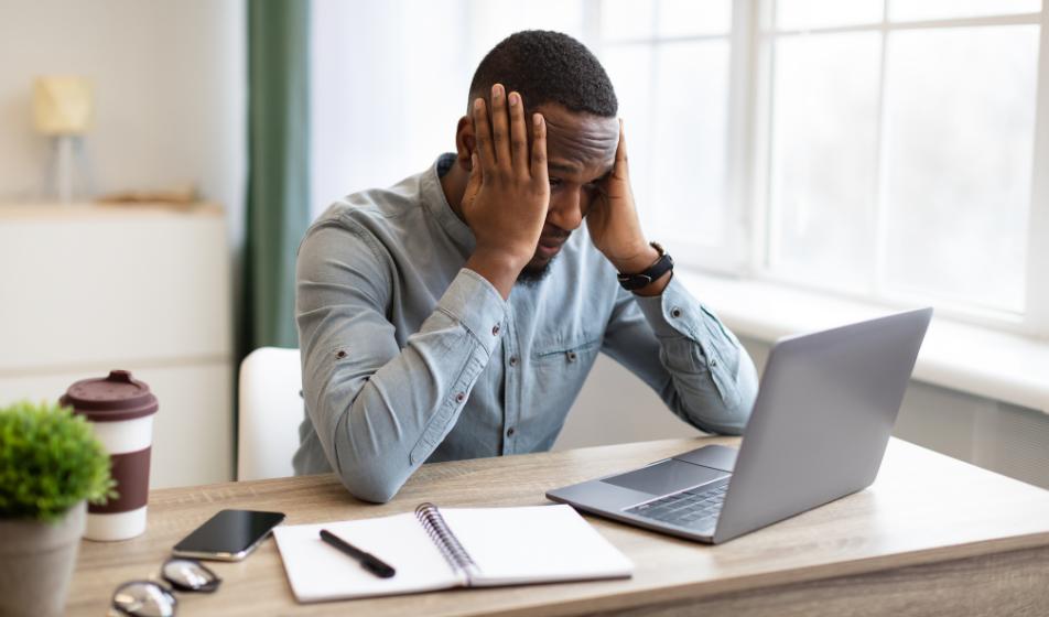 A man in a blue shirt is sat at his desk with his laptop in front of him. He is holding his head in his hands and is looking very stressed.