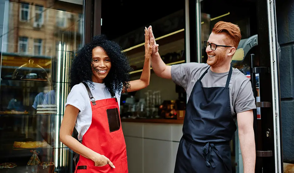 Two baristas are stood outside of a coffee shop and high fiving. One is wearing a bright red apron and the other is wearing a dark blue one.