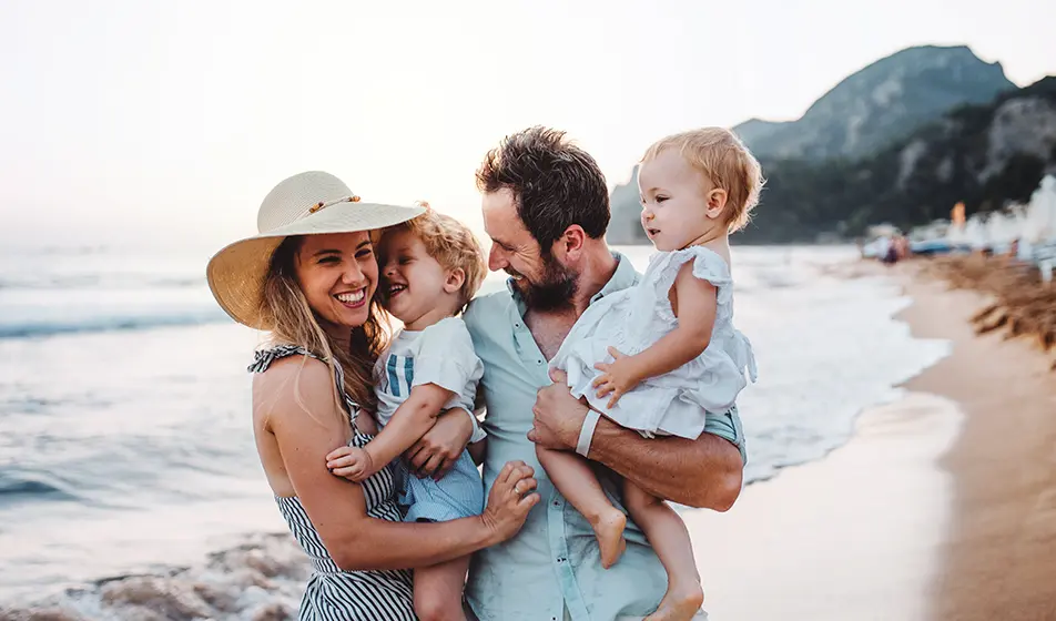 A family of four are on holiday together. They are spending the day at the beach. The mum and dad are holding their toddlers on their hips and everyone is smiling.