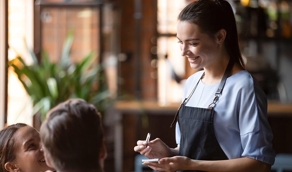 A woman is working at a coffee shop and is taking two customers orders. She is holding a notepad and a pen.
