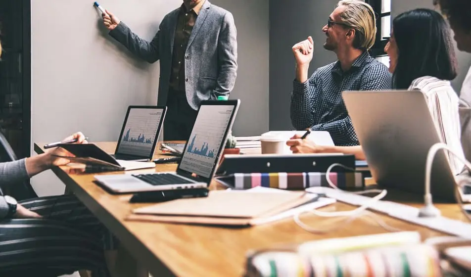 Five colleagues are having a meeting in an office. Four of the colleagues are sat at a wooden desk, listening to their colleague who is stood up. The man stood up is pointing at the wall with a blue pen.