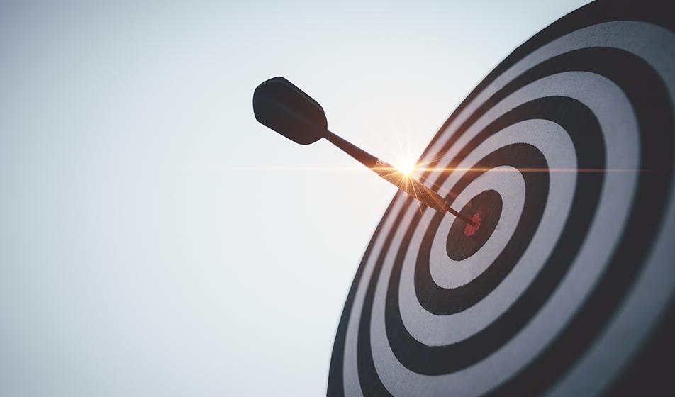 A dart is shown, having hit the centre target on a dartboard. The angle is shown from below at an angle, with the sun in the background.