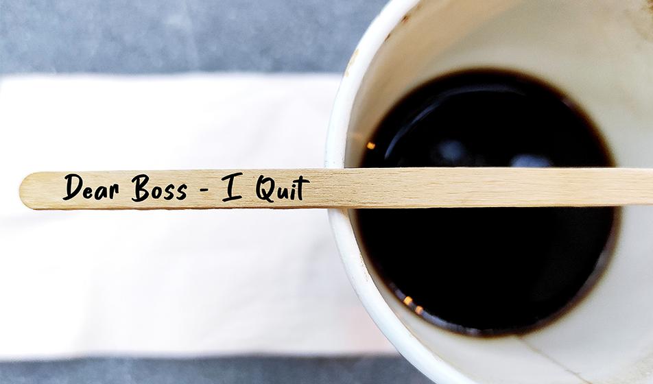A black coffee in a cup is shown. A stirrer stick rests on the cup with 'dear boss - I quit' written on it,
