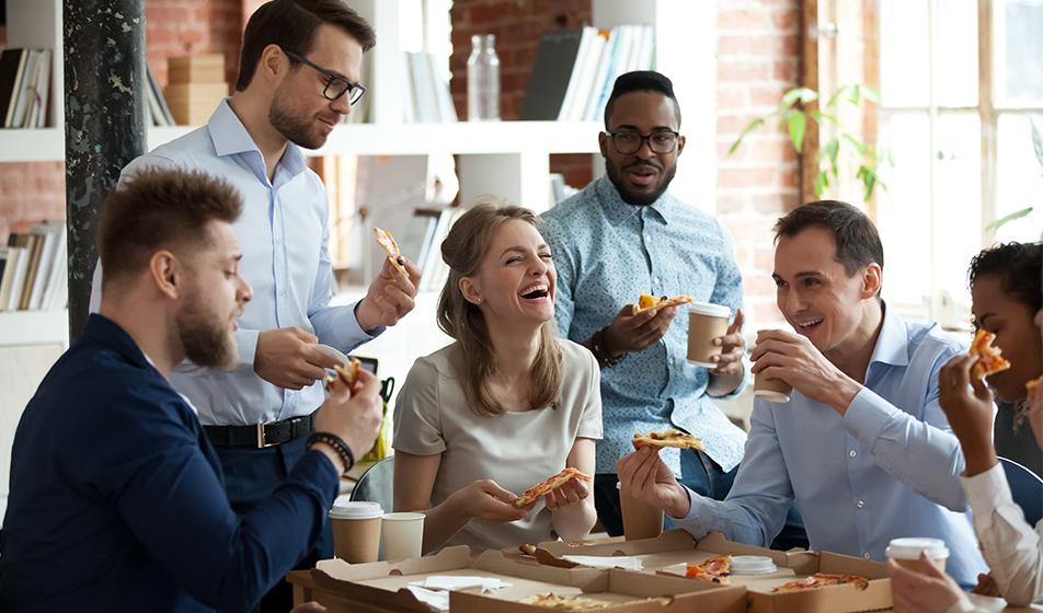 A group of people in an office, laughing, eating pizza out of a box and some holding coffee cups