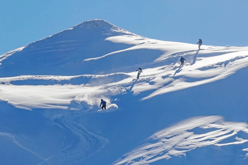 Four people holding poles, skiing off-piste down a wavy path on the side of a snowy mountain.