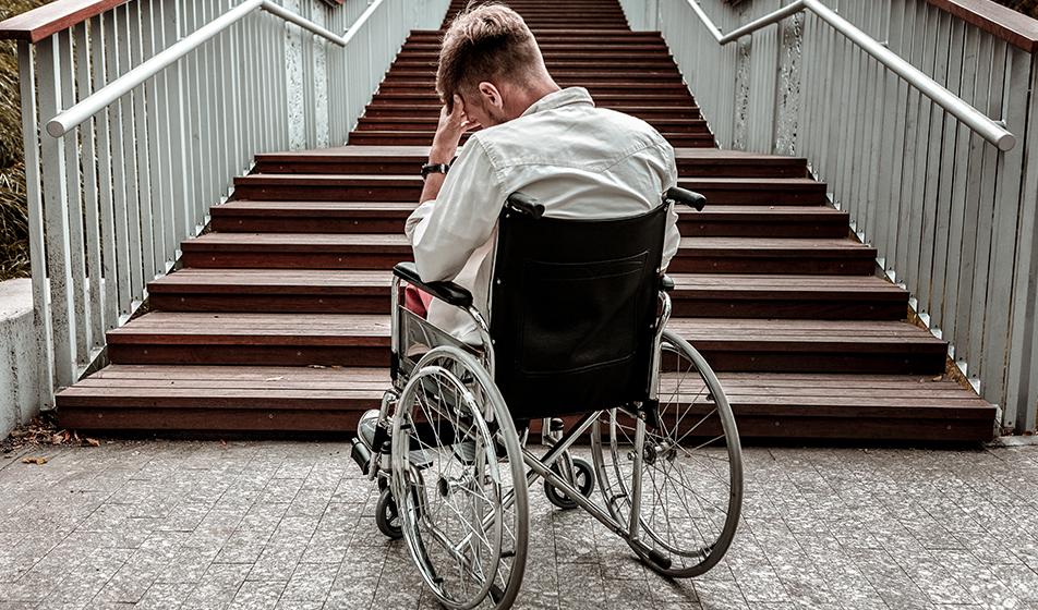 A man in a wheelchair is at the bottom of a long flight of stairs. He is facing away from the camera, with his head in his hands in a desolate manner. 