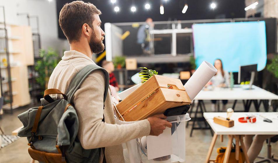 A man with a rucksack on has packed up his desk in a box and is carrying it out, looking sombre.
