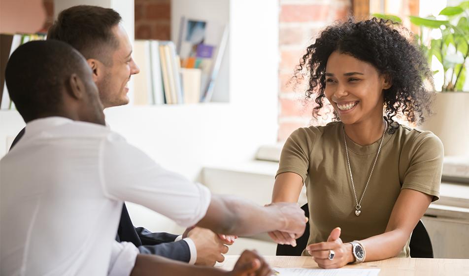 A smiling woman shakes hands across a table with a man. She looks happy. 