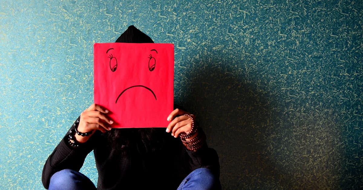 A person is sat on the floor against a blue wall. They are holding a bright red piece of paper over their face which has a sad face on it.