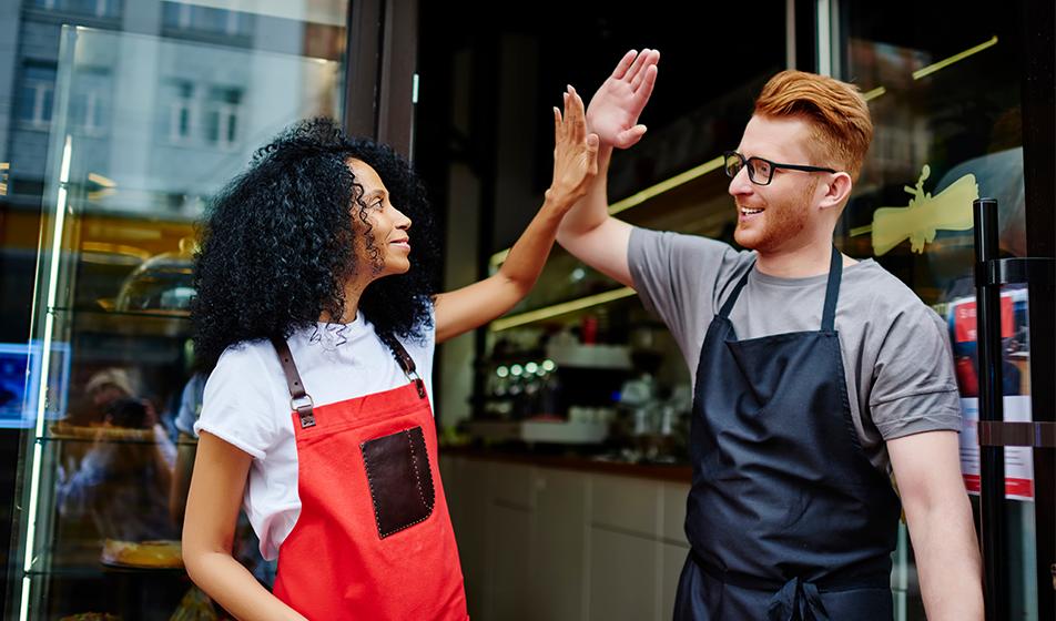 Two hospitality workers are stood outside a coffee shop or restaurant. The woman wears a red apron, the man wears a blue one. They are high-fiving. 