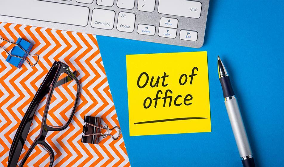 A yellow post-it note reading "out of office" is shown on a blue desk, near a laptop keyboard and some paper clips and a pair of glasses.