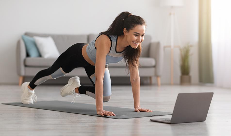 A smiling young woman works out on a mat at home in front of her laptop, she's watching a fitness class.