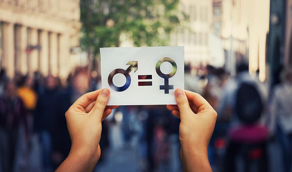 Hands are shown, holding the male and female gender symbols on a white card. In the background, a blurred street with people and buildings out of focus is shown.