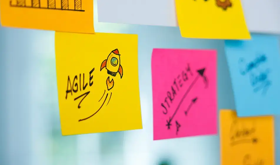 A yellow post-it note saying 'agile' with a hand-drawn rocket next to it is shown stuck on a glass wall, next to a pink post-it note saying 'strategy'