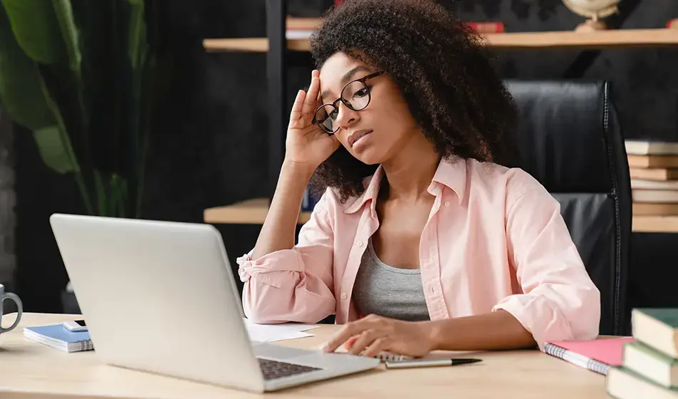 A woman sits at her desk, looking at her laptop, looking very tired and fed up. She rests her head on her hand slightly. 