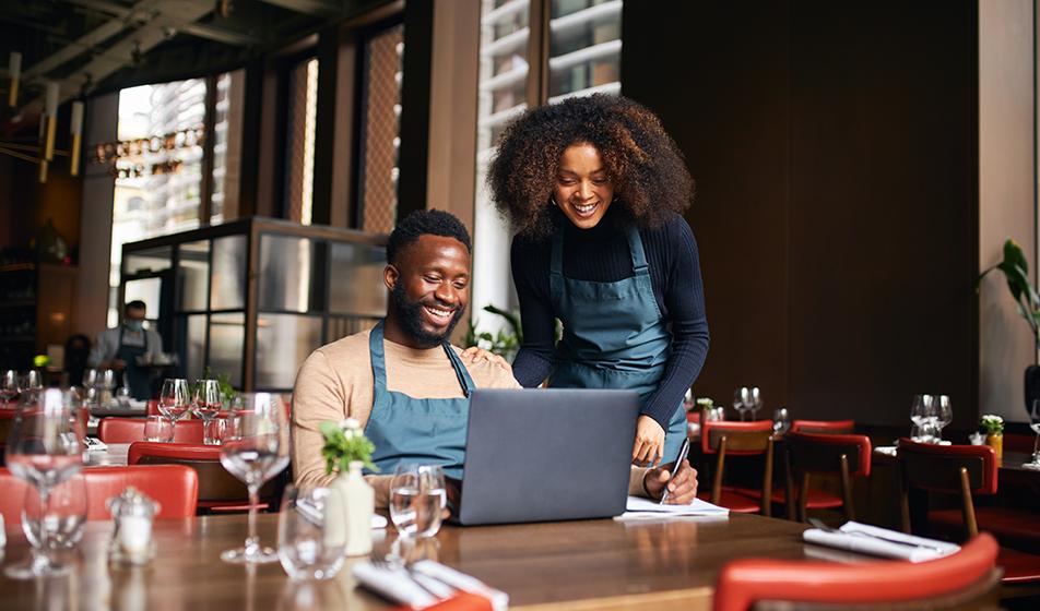 Two restaurant managers look at a laptop in a restaurant. One is sitting, the other standing. Both are smiling. 