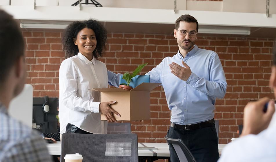 A man stands at the end of a table where people are seated, introducing a woman who appears to be a new employee, holding a box of desk supplies. 