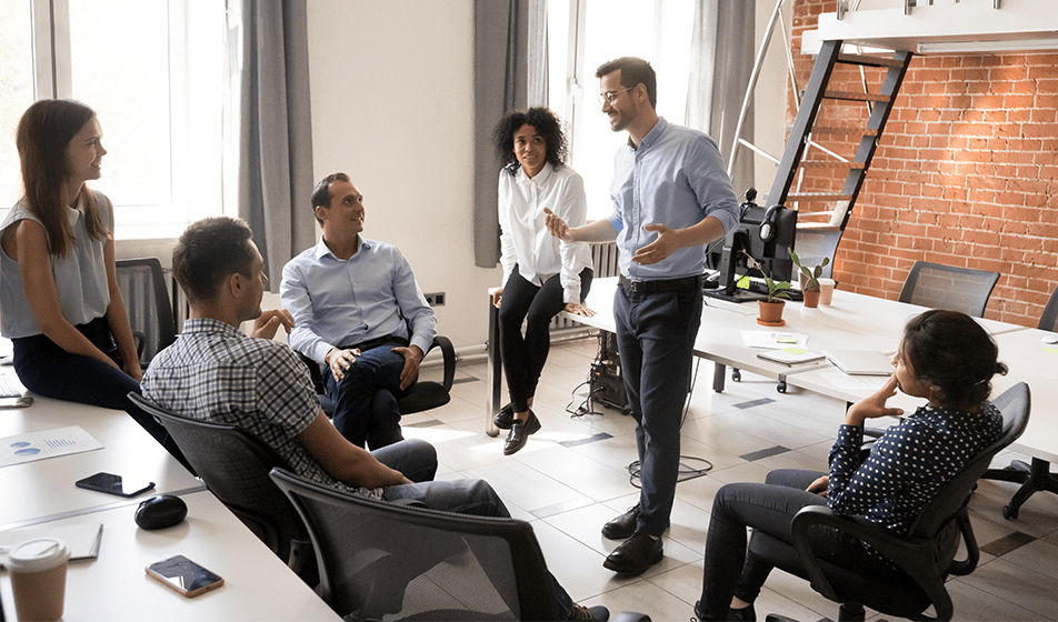 A group of six colleagues are having a relaxed meeting in their modern office. A male colleague is stood up and talking to everyone else who is sat down. They are all smiling at him and engaging with what he is saying. 