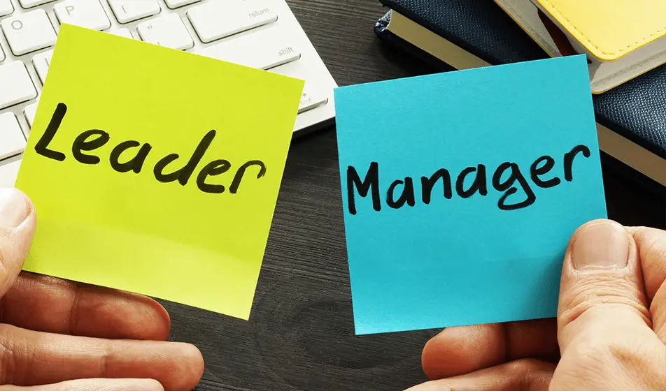 A person is holding two sticky notes side by side. The left hand is holding the yellow sticky note which has the word "Leader" written on it in black ink. The right hand is holding the blue sticky note which has the word "Manager" written on it also in black ink.