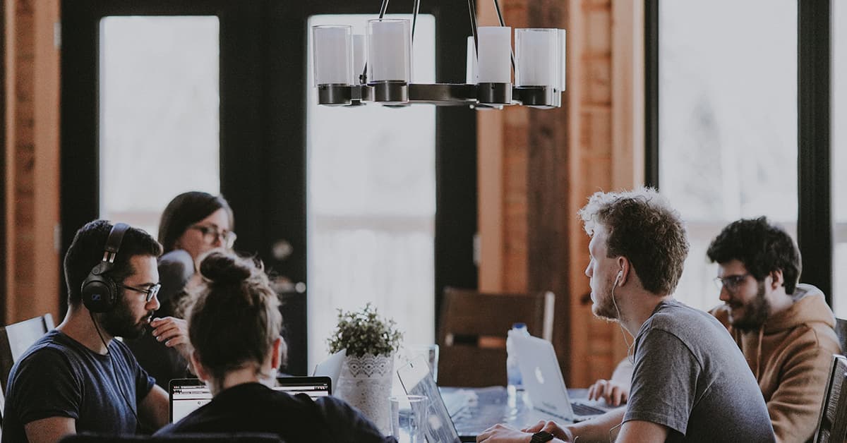 Five people working at a table together. All of them are working on laptops with a small plant in the centre of the table. Above them, there is a modern chandelier hanging from the ceiling.
