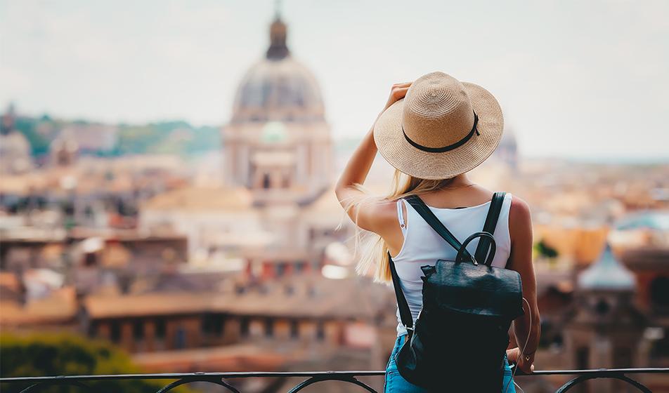 A woman, a tourist with a backpack, stares out at what appears to be the architecture of Rome & the Vatican City. She's seen from behind, wearing a hat, with one hand holding onto her hat. The city is out of focus. 