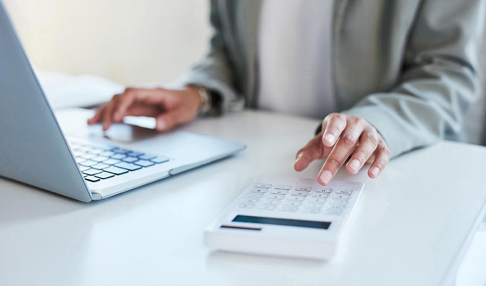 Someone is at a desk with one hand on a laptop and with one hand typing into a calculator. Their hands are the focus of the image. 