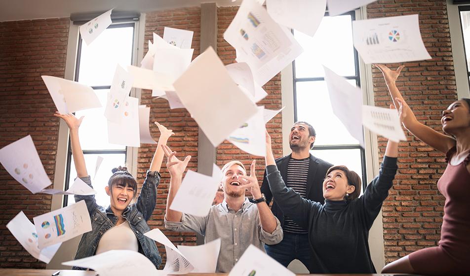 A group of happy employees in an office environment throw lots of paperwork in the air, smiling and laughing. 