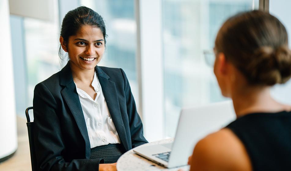 Two women sit across from each other in an office environment. The woman facing the camera is smiling. 