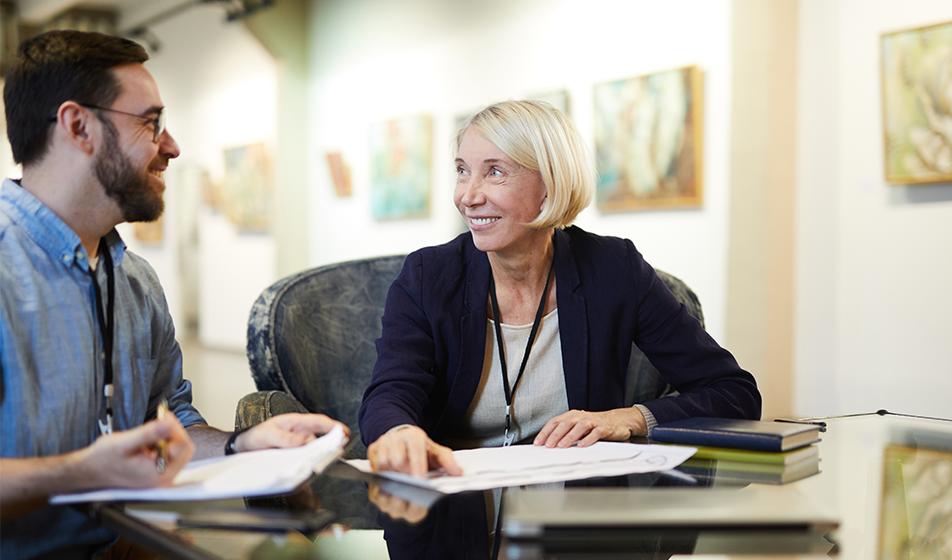 A man and a woman are at a desk in an office, looking at some paperwork in an appraisal environment. The woman is smiling. 