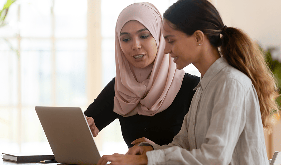 Focused female in a headscarf is mentoring intern worker who is looking at a laptop. Appears to be learning new skills, the mentor is explaining something as they work together at a modern workplace
