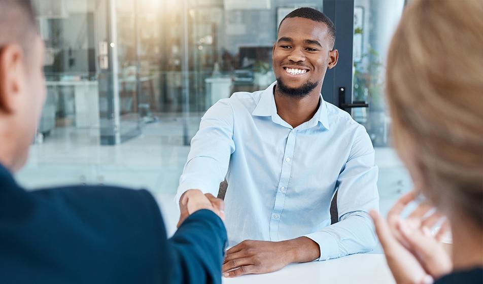 A man sits across a table from a man and a woman in a job interview, shaking the man's hand. He is smiling. 