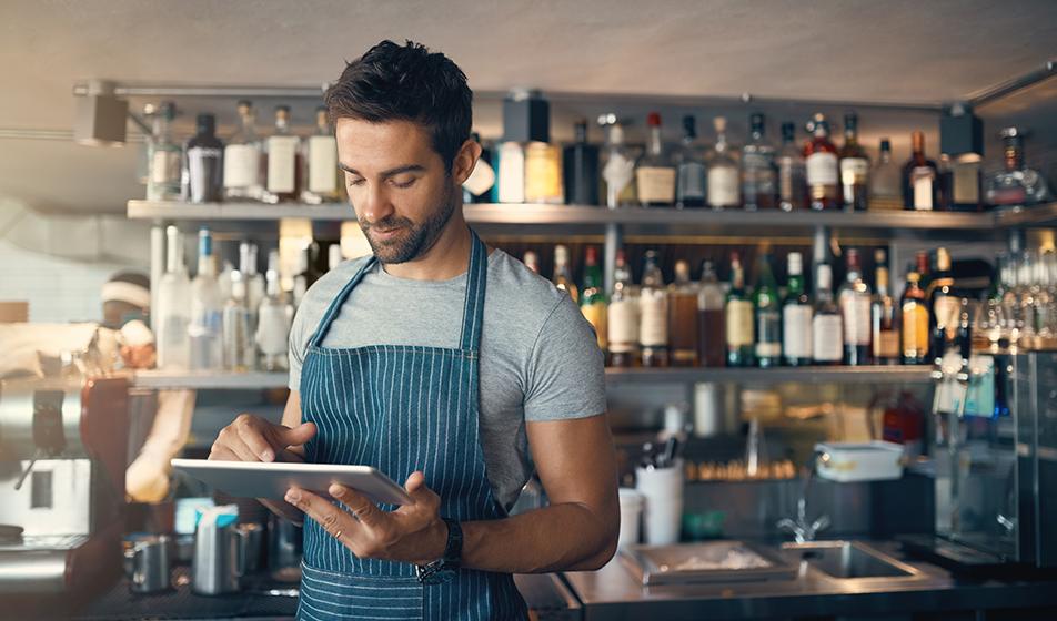 A man in an apron is working his shift in hospitality. He is looking at a digital tablet in his hands. 