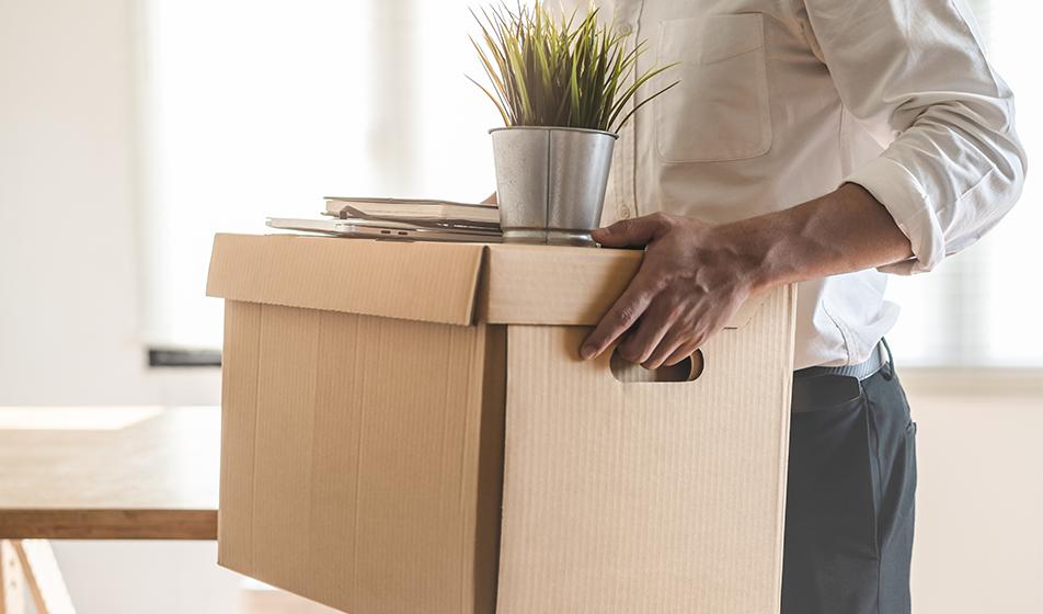 A man is shown holding a cardboard box containing a plant protruding from the top. He's packed up his desk after losing or leaving a job. 