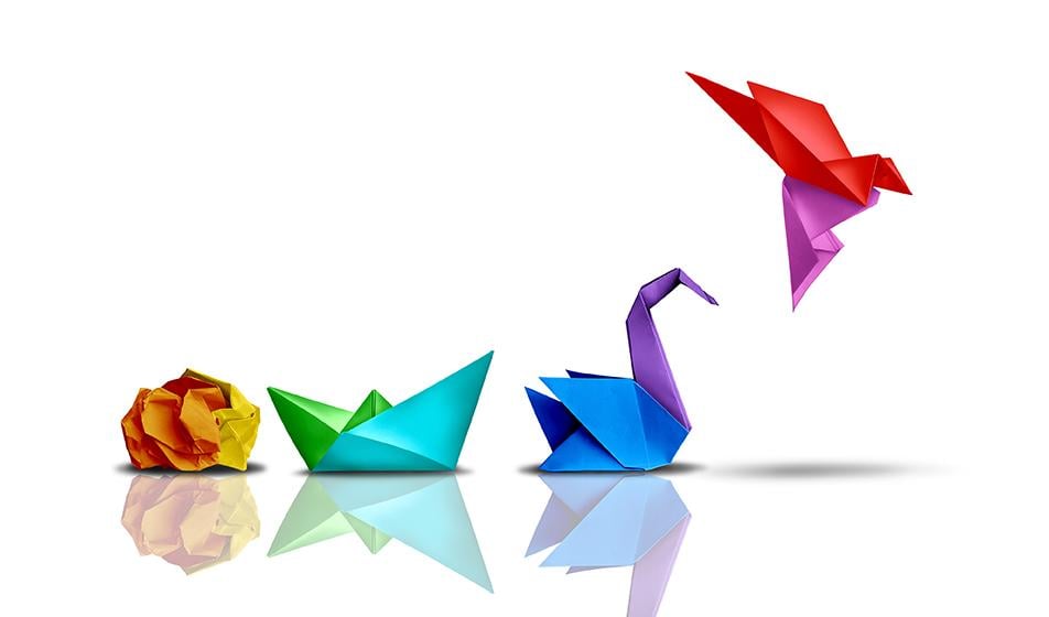Four origami figures are shown, made of different coloured paper. One is yellow and scrunched into a ball, the next is a small turquoise boat, the next is a blue and purple swan, and the final one in the air is a red and pink bird. These represent growth or development
