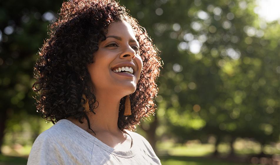 A woman is smiling in the sunshine, with trees in the background.