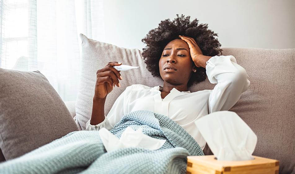 A woman lies on a sofa under a blanket, with tissues nearby. She's looking at a thermometer and looks stressed and unwell. 