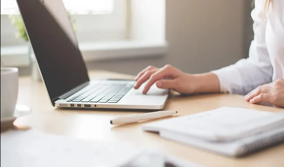 A woman in a white shirt is working at a desk on her laptop. She has her pen and notebook beside her with a white cup.