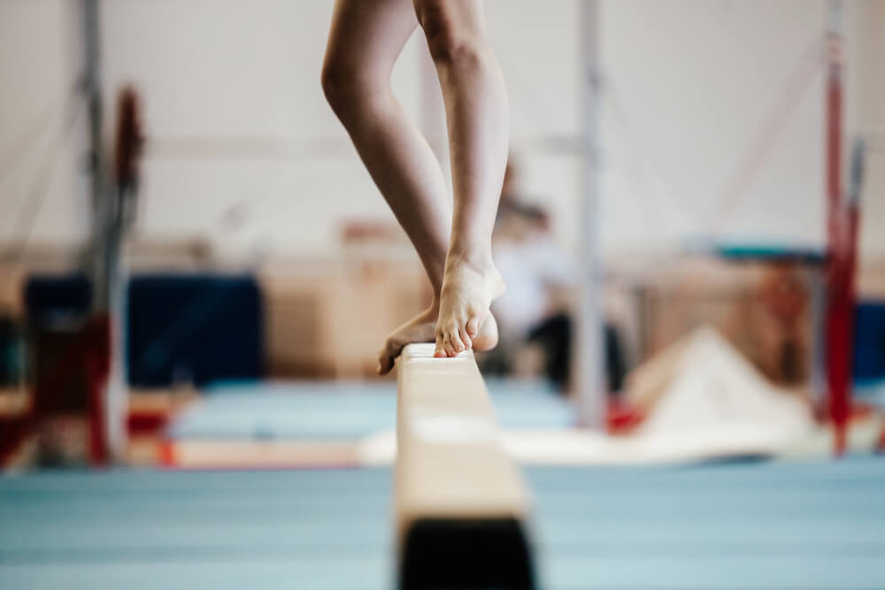 A gymnast is in a gym and walking carefully across a balance beam towards the camera.