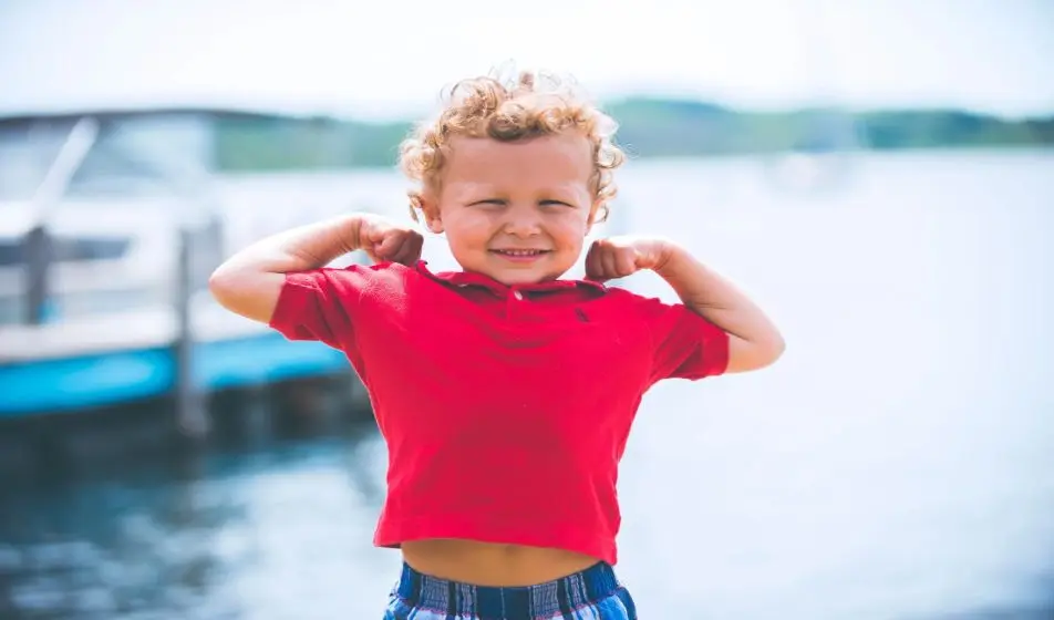 A little boy with curly hair is wearing a bright red t-shirt and blue checkered trousers. He is smiling at the camera and flexing his arms to show off his muscles. Behind him there is the sea and a boat.