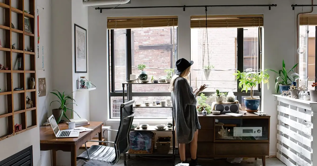 A woman is working from home and is stood up near her window with her phone in her hand. Her laptop is sat on her desk alongside some books and a cup. The room she is working in is full of plants and ornaments spread around the space.