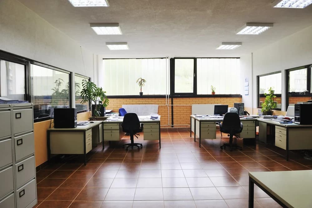 An image of an empty office with a brown tiled floor that has several cream desk spaces within it. Within the office there are several plants spread around the room.