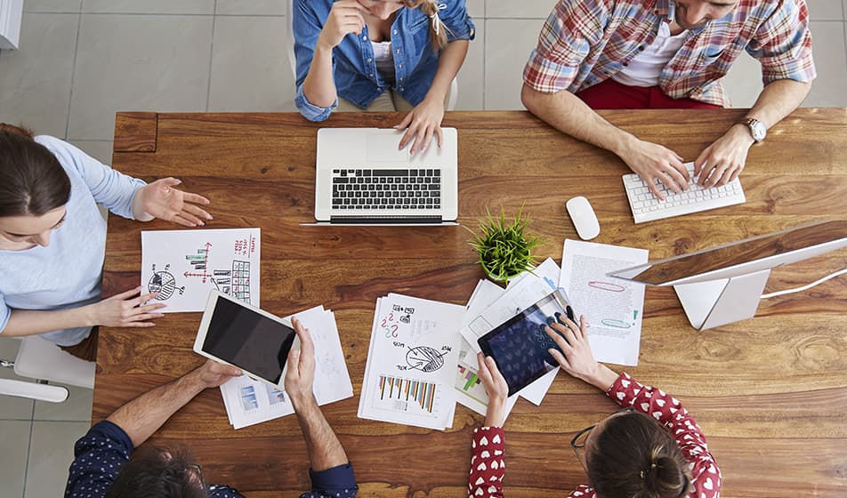 An aerial view of a table in an office environment, where people sit around it with laptops and various pieces of paper in a collaborative meeting. 