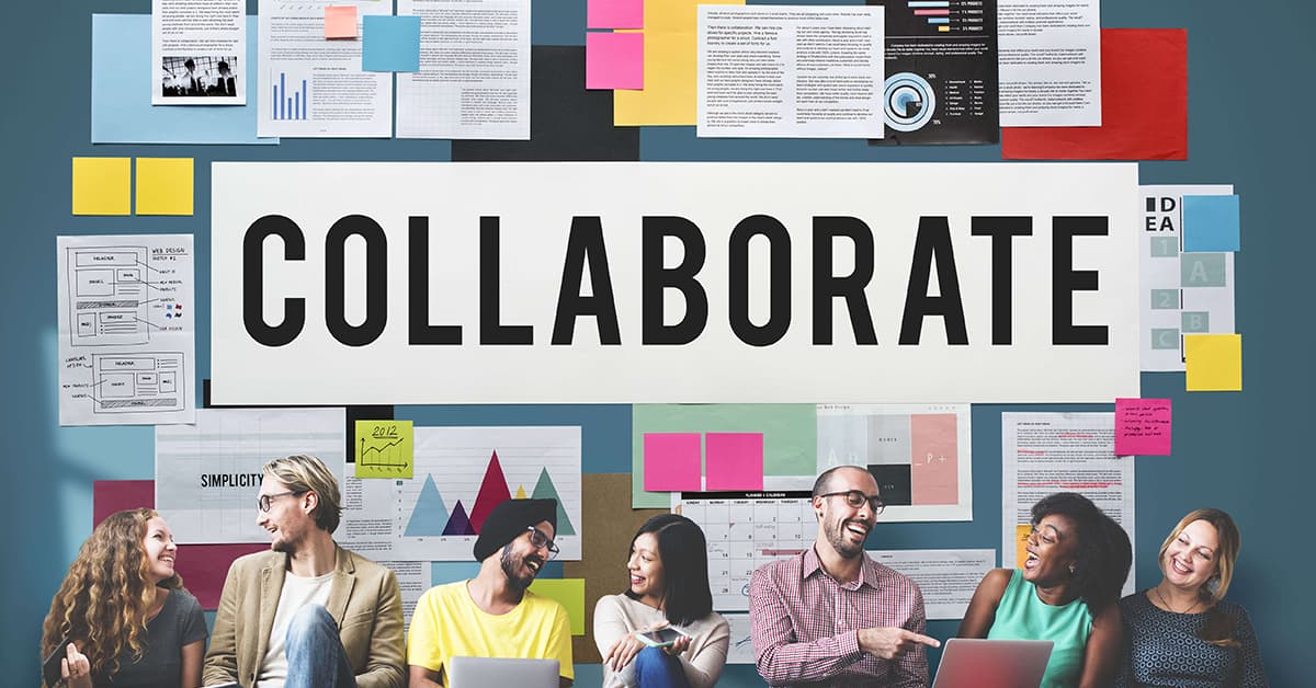 Seven colleagues sat in front of blue wall that is full of different posters and paperwork. In the middle of the wall is the word "collaborate" in large black letters.