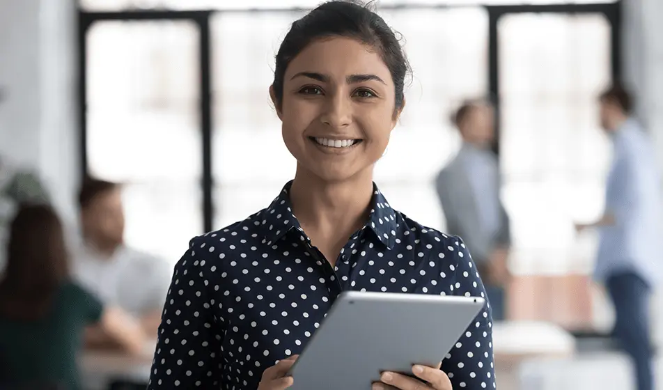 A woman is wearing a dark blue blouse that has white polka dots on. She is holding a tablet and smiling at the camera. In the background, her colleagues are working.