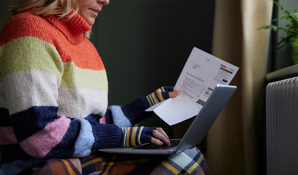 A woman is shown in a thick jumper, fingerless gloves and a blanket, at her laptop by a radiator. She's trying to keep warm, with a paper bill in one hand. 