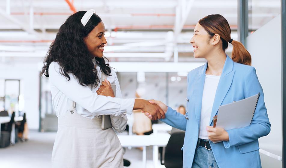 Two women shake hands in an office, smiling