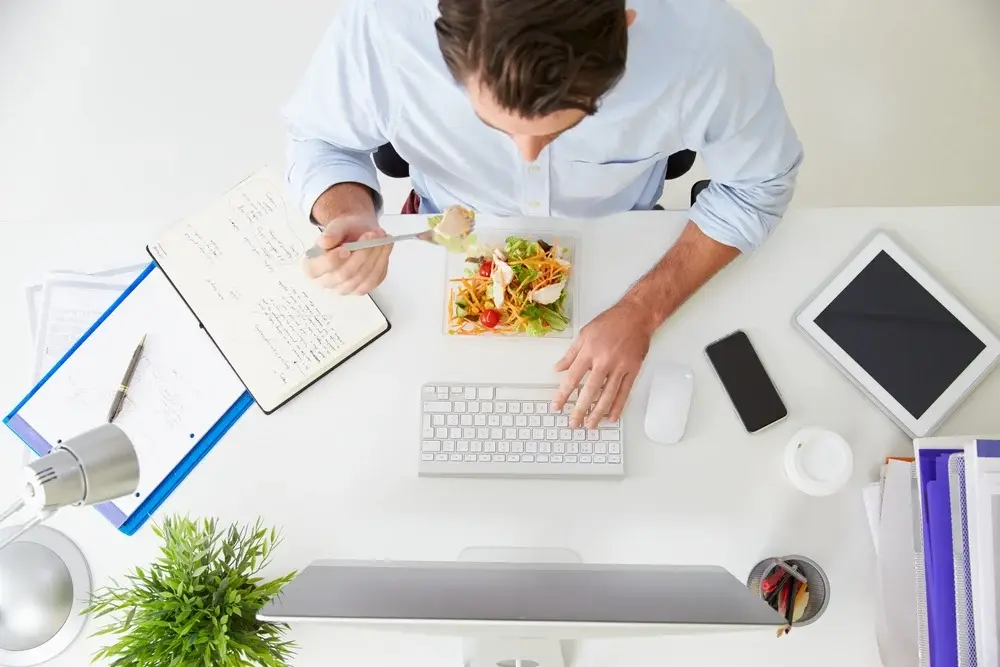 A man is sat at a white desk and eating a salad whilst typing on keyboard. On his desk he has his notebook, a clipboard, his phone, a coffee cup, and a white tablet.