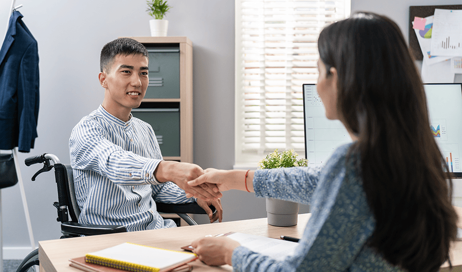 A man in a wheelchair is wearing a striped shirt and is shaking the hand of a woman wearing a blue flowery blouse. This workplace are striving to be inclusive and provide equal opportunities. Everyone has an equal right to employment but disabled people are still under-represented in the UK workforce.